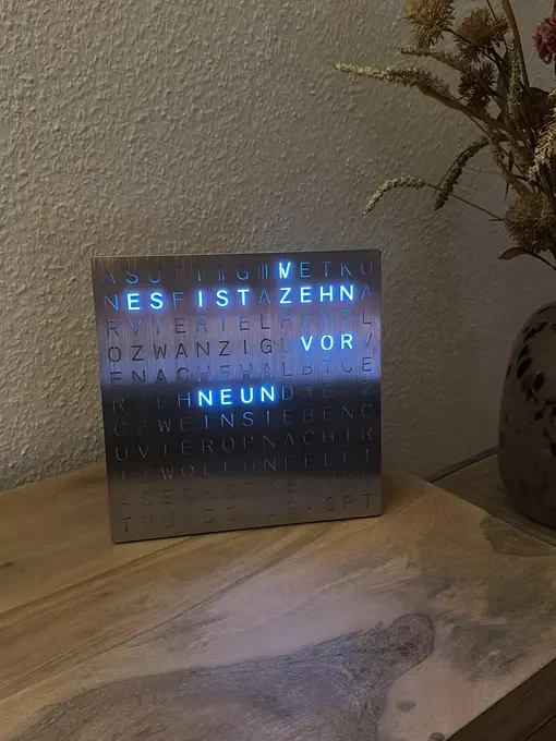Gebürstete Edelstahl-Wortuhr mit blauer Beleuchtung auf einer Holzkonsole