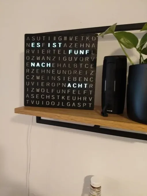 Schwarze Wortuhr auf einem Holzregal neben einer Pflanze