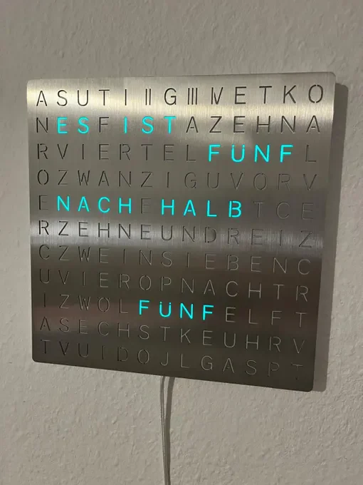 Gebürstete Edelstahl-Wortuhr mit türkis beleuchteten Wörtern an einer weißen Wand
