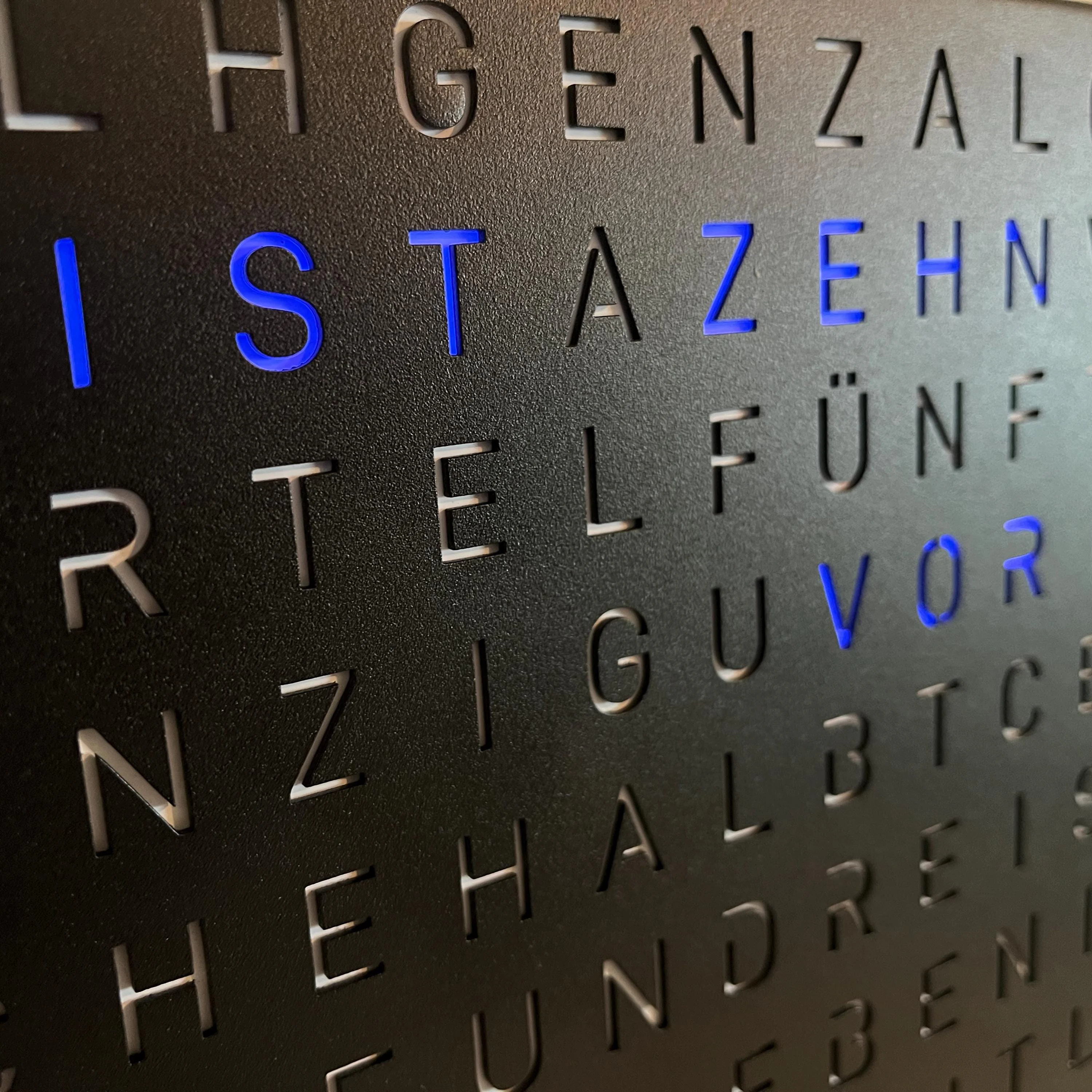 Blau leuchtende Buchstaben der schwarzen TEXTime Wortuhr in Nahaufnahme